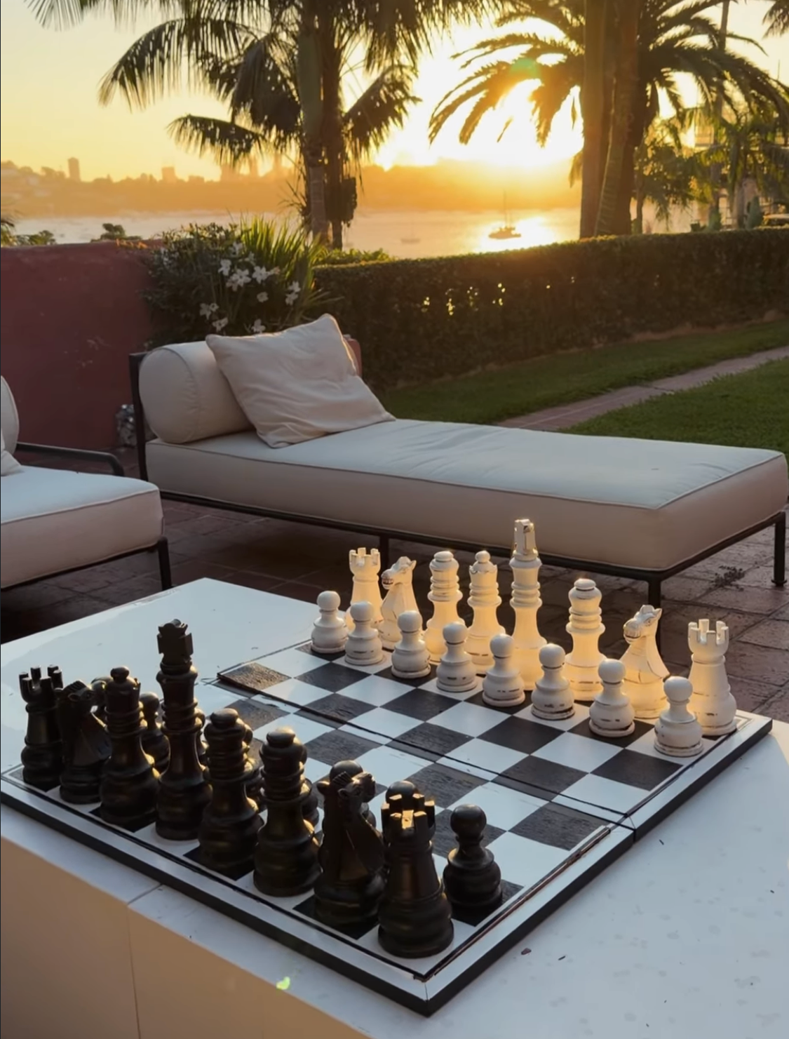 Giant chess set on poolside terrace at golden sunset with cream daybeds, palms and harbour views