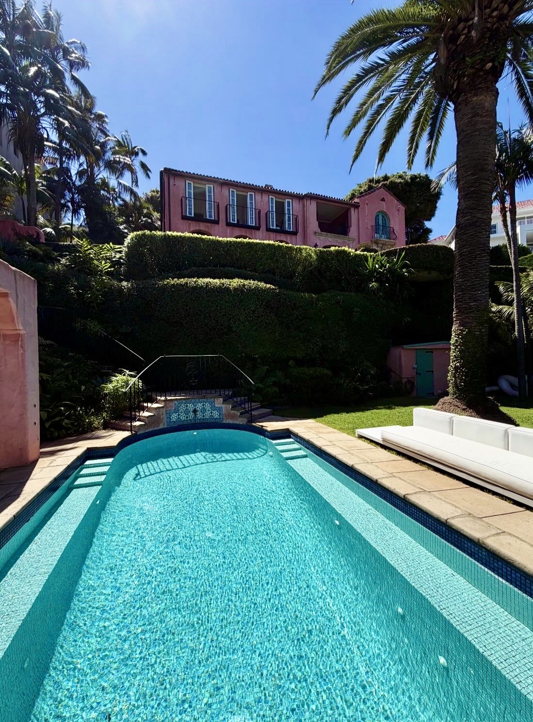 Turquoise lap pool looking back toward Villa Biscaya pink facade, manicured hedges, palm tree and white daybeds under a clear blue sky