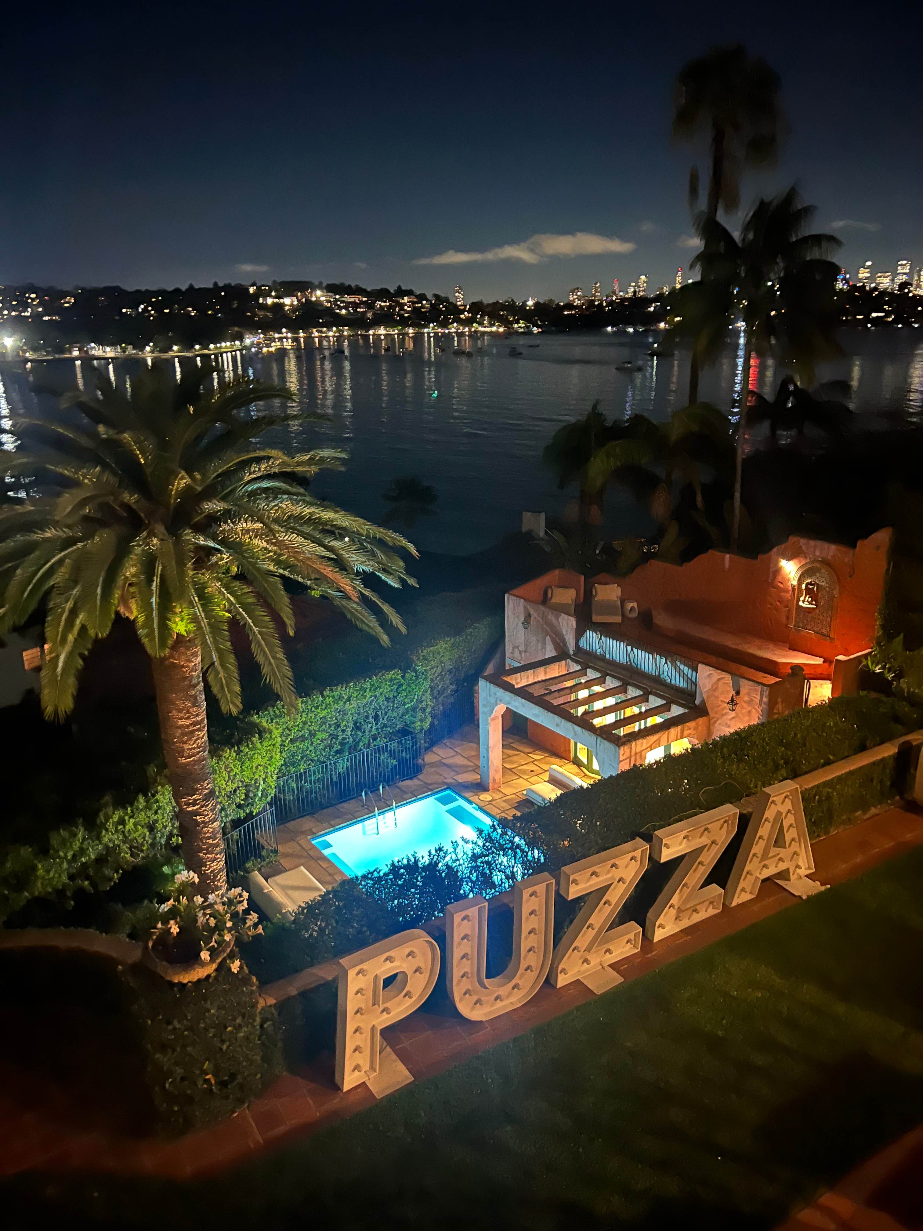 Illuminated marquee letter lights beside the lit turquoise plunge pool at night with the Sydney skyline beyond