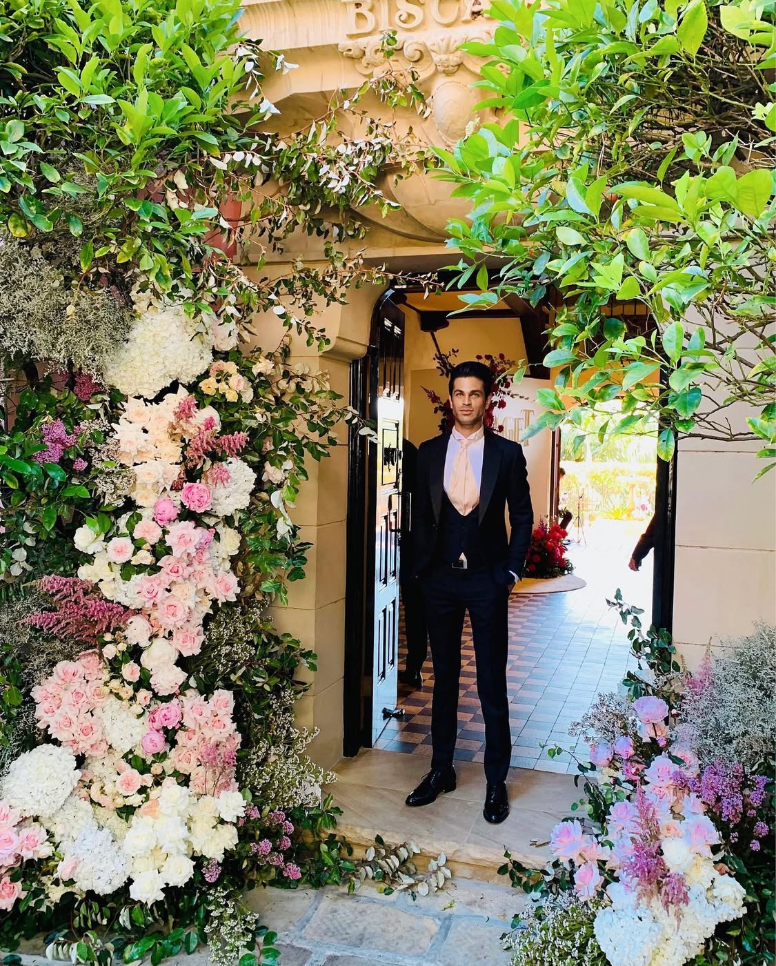 Groom in black tuxedo with peach tie at Villa Biscaya entrance under the carved sandstone Biscaya arch, framed by a lush floral installation of pink roses, white hydrangeas and cascading greenery
