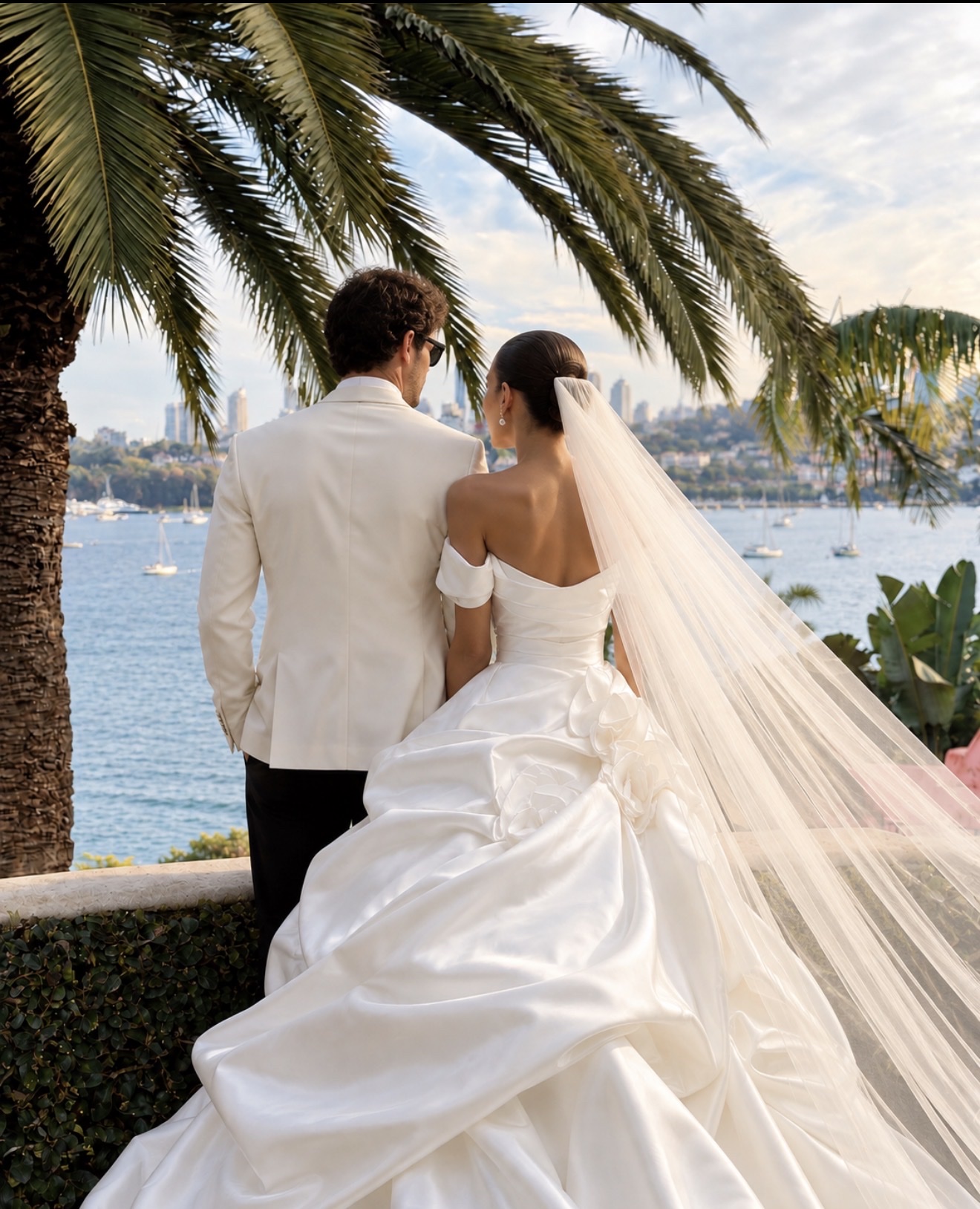 Bride in an off-shoulder white gown with a long veil and groom in an ivory dinner jacket, seen from behind beneath a palm tree looking out over Sydney Harbour