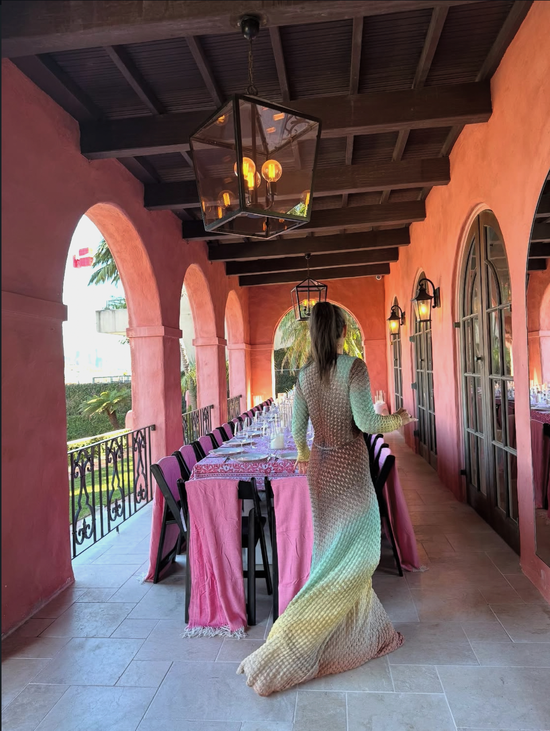 Woman walking through arched terracotta loggia with long banquet tables draped in pink, black chairs, hanging lanterns and harbour views